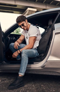 Modern spor arabasında oturan güneş gözlüklü ve günlük kıyafetli yakışıklı bir genç.