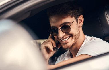 Güneş gözlüklü ve günlük giysileri olan yakışıklı genç adam modern spor arabasında oturuyor ve cep telefonuyla konuşurken gülümsüyor.