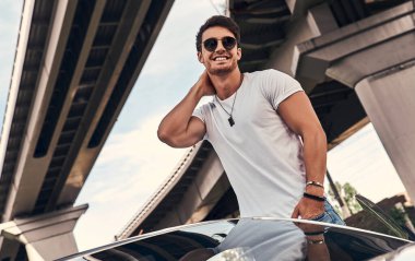 Sokakta modern spor arabasının yanında duran, günlük kıyafetli yakışıklı genç adam.