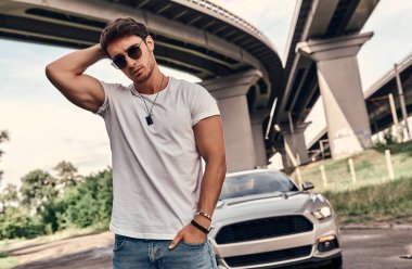 Sokakta modern spor arabasının yanında duran, günlük kıyafetli yakışıklı genç adam.