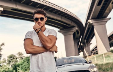 Sokakta modern spor arabasının yanında duran, günlük kıyafetli yakışıklı genç adam.
