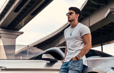 Sokakta modern spor arabasının yanında duran, günlük kıyafetli yakışıklı genç adam.