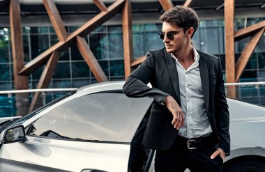 Takım elbiseli yakışıklı genç işadamı modern iş merkezinin önünde spor arabasının yanında duruyor.