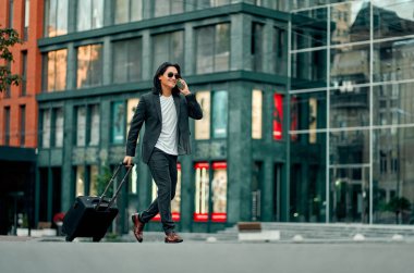 Şehirdeki Asyalı işadamı. Kendine güveni tam genç bir adam takım elbiseli, güneş gözlüklü ve elinde bavulla akıllı bir telefonla konuşuyor.
