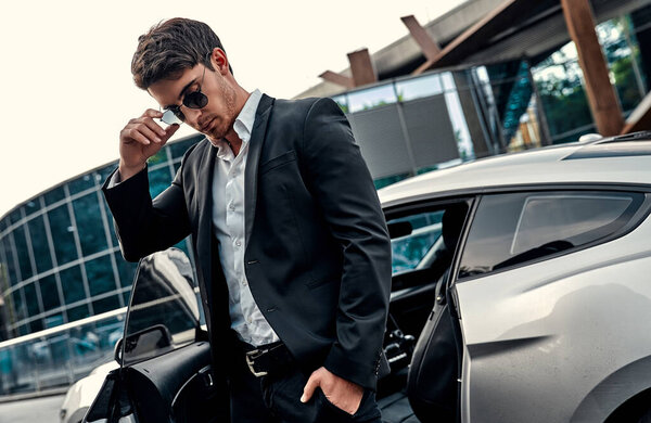Handsome young businessman in suit is standing near his sport car in front of modern business center