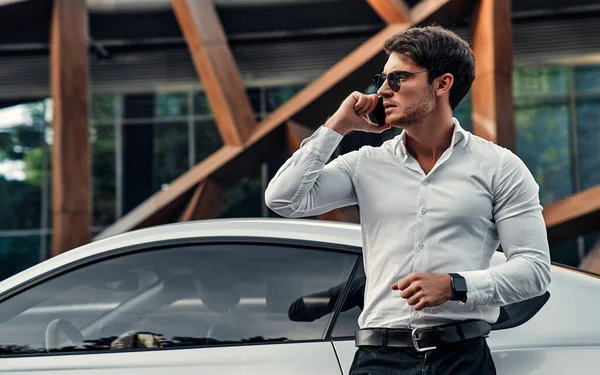 一位穿着西服的英俊的年轻商人站在他的跑车旁边 站在现代商业中心前面 用手机说话 — 图库照片