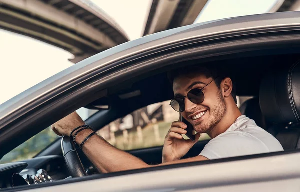 穿着太阳镜和休闲装的英俊年轻人坐在他的现代跑车上 一边笑着一边用手机说话 — 图库照片