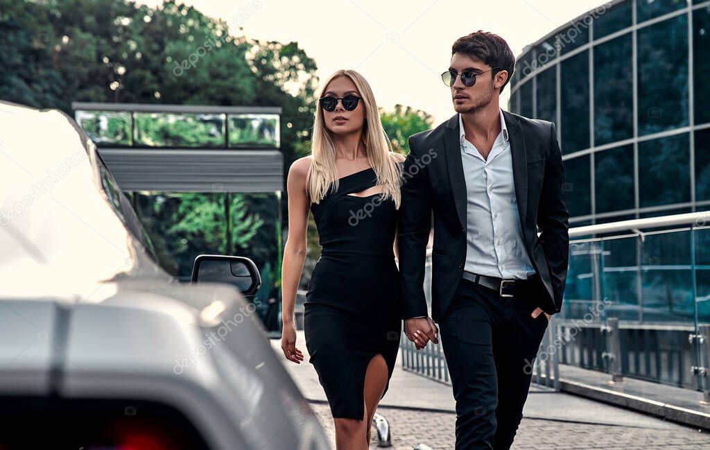 Beautiful young couple near sport car. Attractive blonde in elegant black dress and handsome businessman in suit walking in front of modern business center.