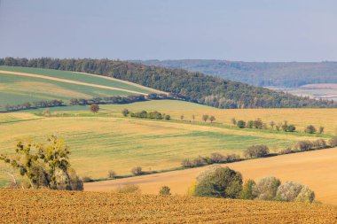 Rolling hills and farmland under a clear sky create a peaceful rural landscape. Textured fields in warm earth tones are visible. Moravia region of Czech Republic, Europe.