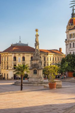 Pecs, Macaristan - 13 Temmuz 2024: Szechenyi Meydanı. Üzerinde veba sütunları olan tarihi bir kare, açık mavi gökyüzü altında klasik mimari ile çevrili..