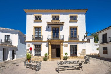 Ronda, İspanya - 4 Eylül 2024: Balkonları ve güneşli bir plazada süslü bankları olan beyaz yıkama binası. Huzurlu, resimli bir Avrupa şehir meydanı.