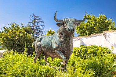 Ronda, İspanya - 4 Eylül 2024: The Plaza de Toros, Ronda 'da bir arenada. Bronz boğa heykeli görkemli bir şekilde yemyeşil, açık havada canlı mavi bir gökyüzünün altında duruyor..