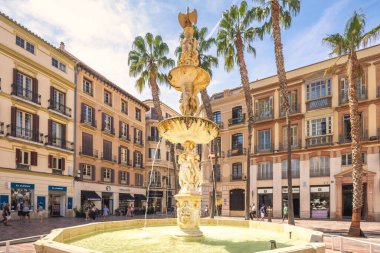 Malaga, İspanya - 12 Eylül 2024: Palmiye ağaçları ve tarihi binalarla çevrili güneşli bir plazada süslü bir çeşme. İnsanlar sıcak havanın tadını çıkararak geziniyor..