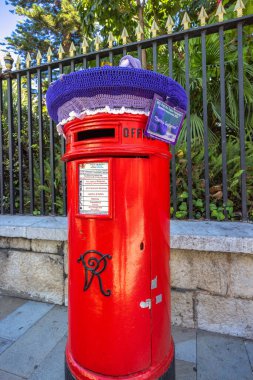 Cebelitarık, Birleşik Krallık - 10 Eylül 2024: standart İngiliz tasarımı Viktorya dönemi posta kutusu. El örgüsü mor ve lavanta şapkalı kırmızı bir posta kutusu. Büyüleyici bir şehir manzarası..