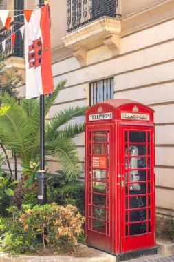 Cebelitarık, Birleşik Krallık - 10 Eylül 2024: Klasik bir kırmızı telefon kulübesi bir bayrak ve yemyeşil bir binanın yanında duruyor. Büyüleyici bir sokak sahnesi.