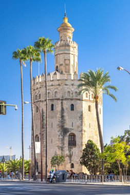 Seville, İspanya - 8 Eylül 2024 Torre del Oro gözlem kulesi. Antik taş kule, palmiye ağaçları ve şehir hayatıyla çevrili canlı mavi gökyüzüne karşı dimdik ayakta duruyor..