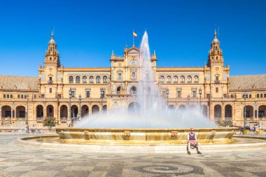 Seville, İspanya - 8 Eylül 2024: İspanya 'nın başkenti Endülüs' teki Plaza de Espana Meydanı. Güneşle sırılsıklam olmuş bir meydanda büyük bir çeşmenin yanında dinlenen kişi, muhteşem bir bina oluşturur..
