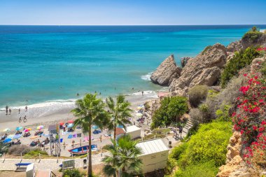 Nerja, İspanya - 9 Eylül 2024: Turkuaz suyu, palmiye ağaçları ve kayalık kayalıkları olan sakin bir sahil koy. İnsanlar kumlu sahilde rahatlar..