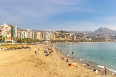 Malaga, İspanya - 12 Eylül 2024: Güneşli bir gökyüzünün altında güneşli banyoları, binaları ve sakin okyanusları olan kumsal.