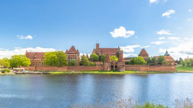 Malbork Kalesi, Kuzey Polonya, Avrupa 'da tuğladan bir Gotik kale. Durgun sulara yansıyan görkemli şato kompleksi, serpilmiş bulutlarla dolu berrak bir gökyüzünün altında yemyeşil tarafından kucaklanıyor..