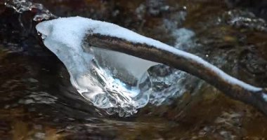 Akan bir derenin bir dalındaki buz oluşumunun yakın çekimi, kışın güzelliğini ve suyun gücünü gözler önüne seriyor. Kar, dalın tepesinde, suyun üstünde..