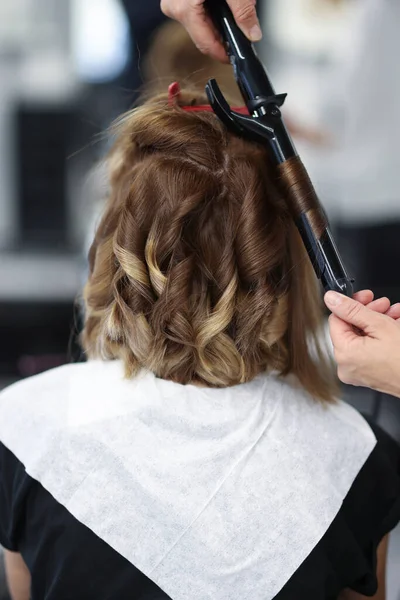 Woman in beauty salon is twisted curls of hair on curling iron. — Stock Photo, Image