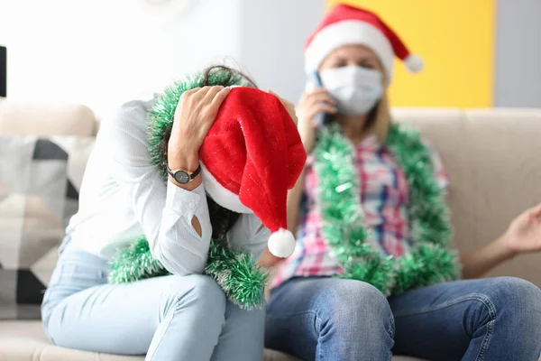 Kvinna i Santa claus hatt sitter på soffan med huvudet böjt bredvid kvinnan i medicinsk mask talar i telefon — Stockfoto