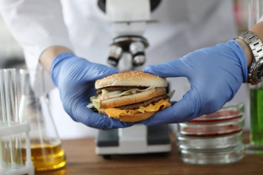 Lastik eldivenli bilim adamları, kimya laboratuvarında mikroskop ve cam test tüplerinin önünde hamburger tutuyor.
