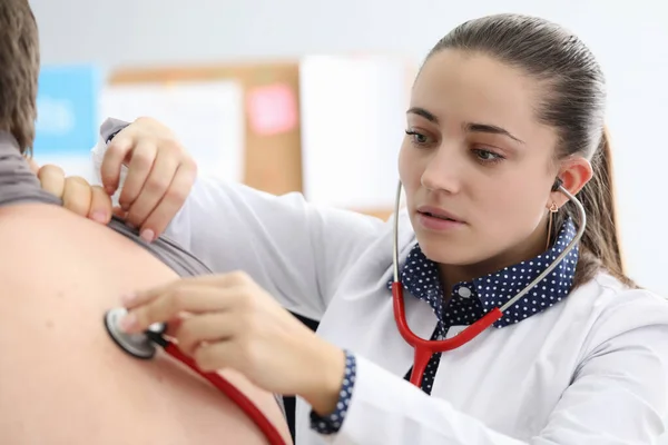 Mujer joven médico escucha los pulmones de paciente con retrato de estetoscopio rojo — Foto de Stock