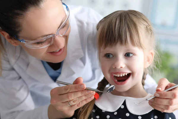 Donna dentista in occhiali protettivi esamina i denti della bambina paziente con strumenti in metallo ritratto — Foto Stock