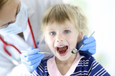 Koruyucu maske takan doktor küçük kızın çenesini ve ağzını muayene ediyor.