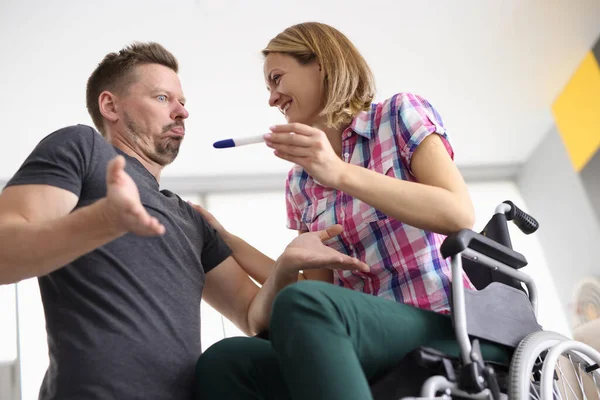 Жінка в інвалідному візку щасливо посміхається чоловікові і тримає тест на вагітність чоловік виглядає здивованим . — стокове фото