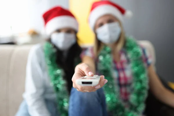Deux copines sont assises sur un canapé avec télécommande en chapeaux Santa Claus et masques médicaux de protection. — Photo