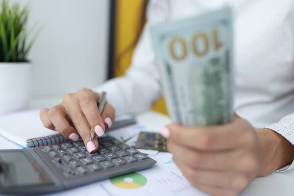 Female hands are holding one hundred dollar bills and working on calculator.