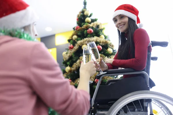 Жінка-інвалід з червоним капелюхом Санти на голові в інвалідному візку п'є шампанське з другом біля ялинки вдома — стокове фото