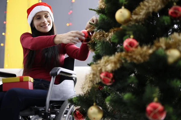 Молода жінка сидить у інвалідному кріслі в червоному капелюсі Санти Клауса і прикрашає ялинку іграшками — стокове фото