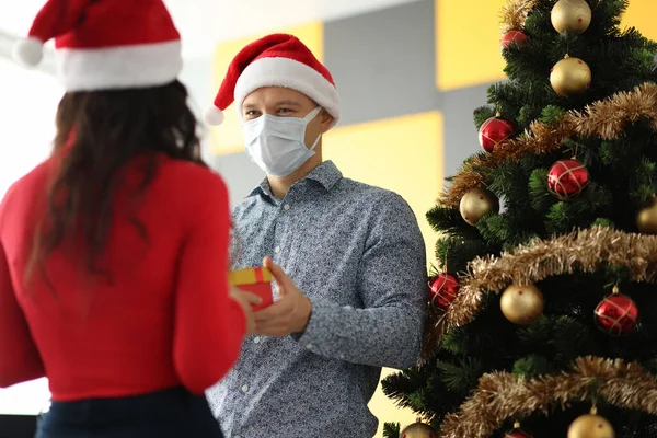 Hombre joven con sombrero de santa claus y máscara protectora en la cara dando regalo a la mujer cerca del árbol de Navidad — Foto de Stock