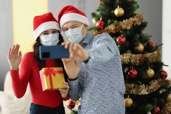 Homme et femme en chapeaux de Père Noël et masques de protection sur les visages tiennent un téléphone portable et agitent près de l'arbre de Noël à la maison — Photo