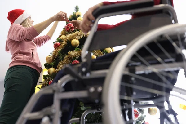 Frau schmückt Weihnachtsbaum neben Mann im Rollstuhl. — Stockfoto