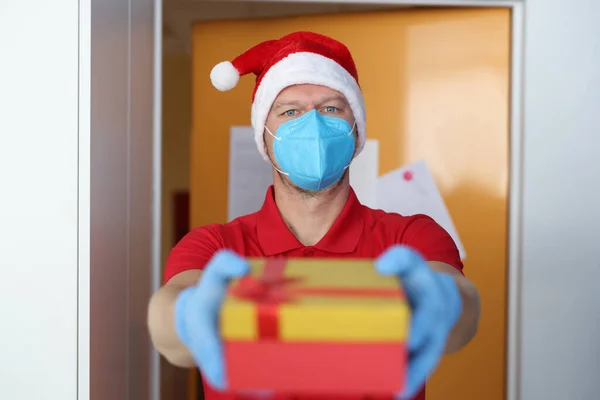 Mensageiro masculino usando máscara protetora médica e chapéu de Papai Noel segura uma caixa acessível. — Fotografia de Stock