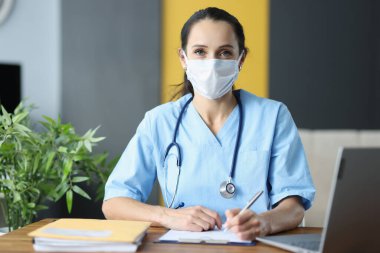 Koruyucu tıbbi maskeli bir doktor ofisinde oturuyor ve belgeleri yazıyor.