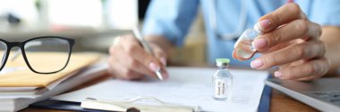 Doktor masada doküman yazıyor ve Coronavirus enfeksiyonuna karşı aşıyla şişeyi yakın plan tutuyor.