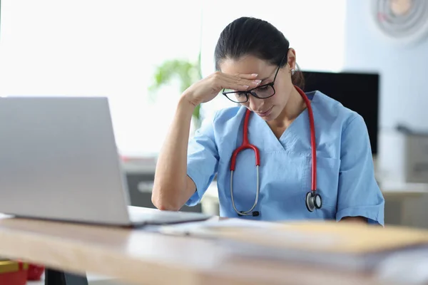 Ung kvinna läkare sitter vid bordet och håller huvudet framför laptop på kontoret — Stockfoto