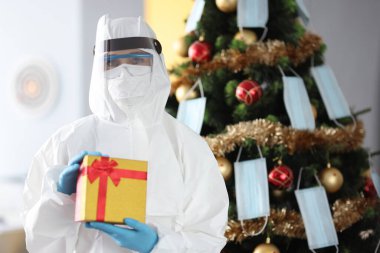 Koruyucu kıyafetli doktor Noel ağacının yanında duruyor ve hediyesini tutuyor.