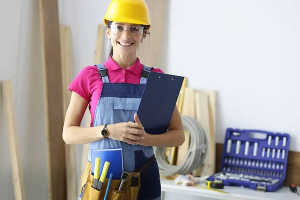 Jonge vrouw bouwer in beschermende overalls en harde hoed glimlachen in de werkplaats — Stockfoto