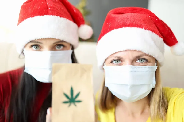 Dos mujeres con sombreros de Santa Claus y máscaras protectoras en rostros sosteniendo bolsa de marihuana — Foto de Stock