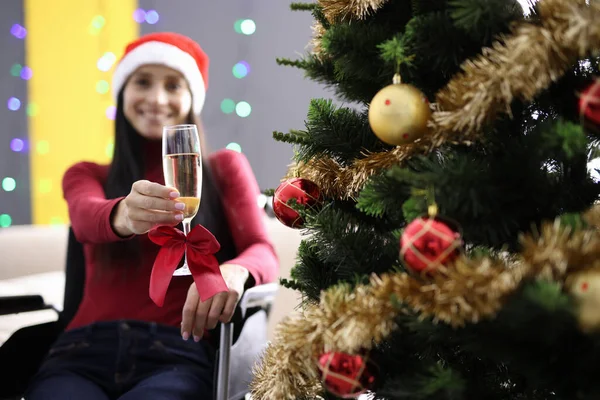 Smiling woman in santa hat sitting in wheelchair and holding glass of champagne