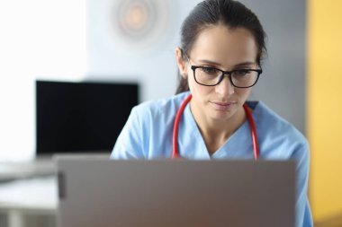 Gözlüklü genç bayan doktor klinikte dizüstü bilgisayarın başında oturuyor.