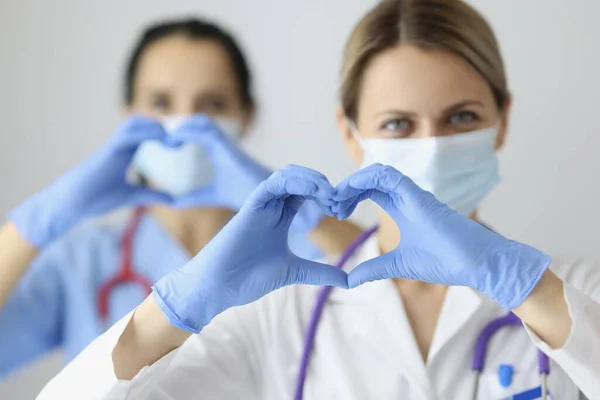 Dos doctores en máscaras médicas y guantes de goma mostrando el corazón con las manos —  Fotos de Stock