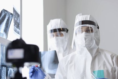 Koruyucu ekranlar ve veba önleyici giysiler giyen doktorlar röntgen odasındaki kameraya bakıyor.